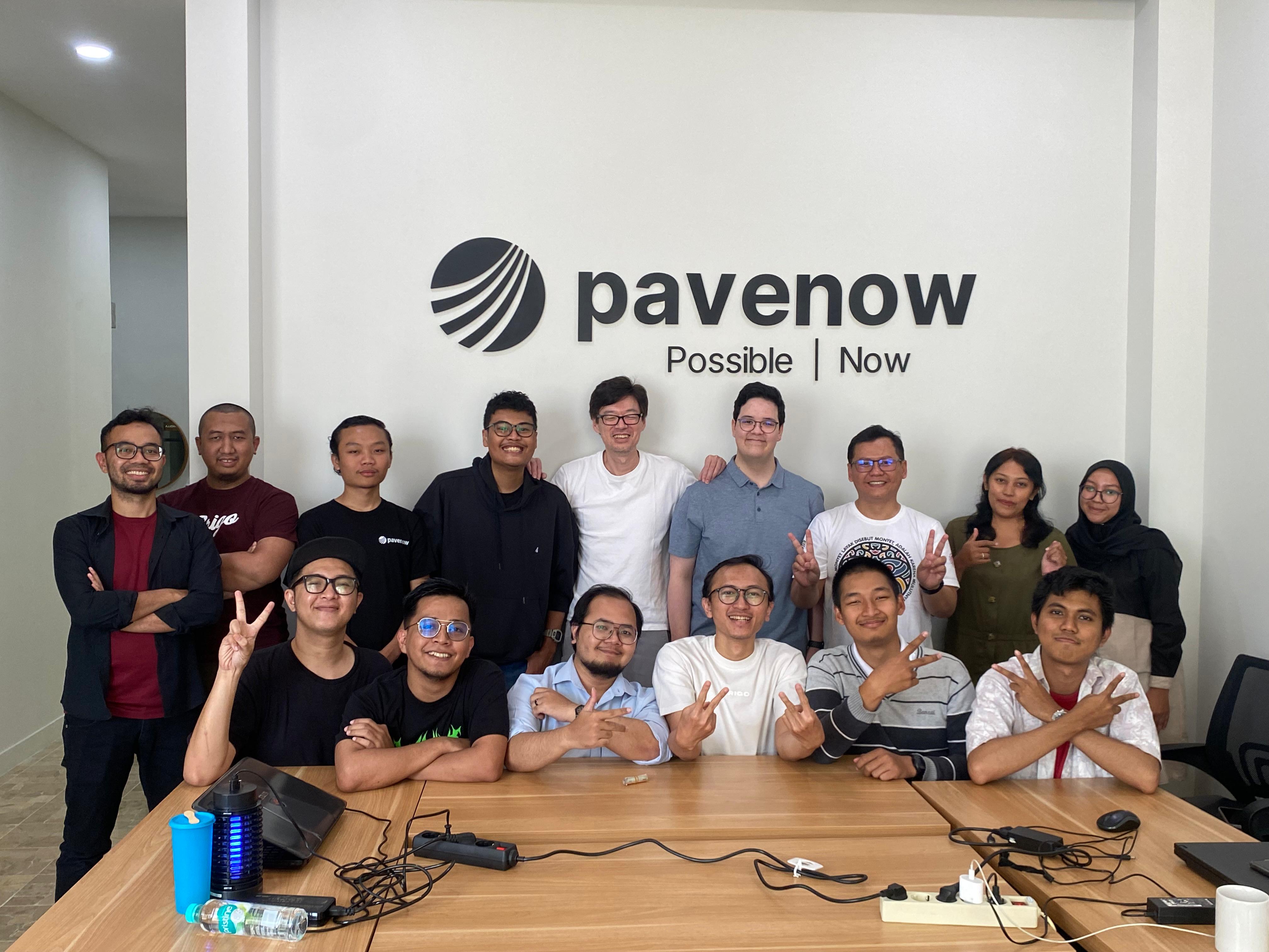 Picture of the Pavenow team working together in Bandung. A group of people sitting around a table, with laptops open and drinks on the table. The room is well-lit and has a modern, cozy feel.